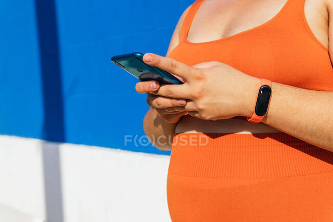 Anonimo plus size atleta femminile etnica in abbigliamento attivo con cellulare contro la parete piastrellata blu alla luce del sole — Foto stock