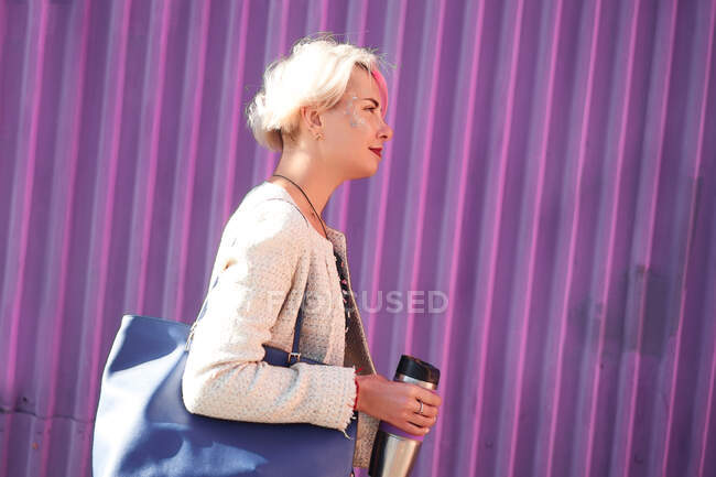 Vue latérale d'une femelle informelle positive avec des cheveux courts teints debout avec une boisson dans une tasse écologique sur fond de mur violet en ville et regardant ailleurs — Photo de stock