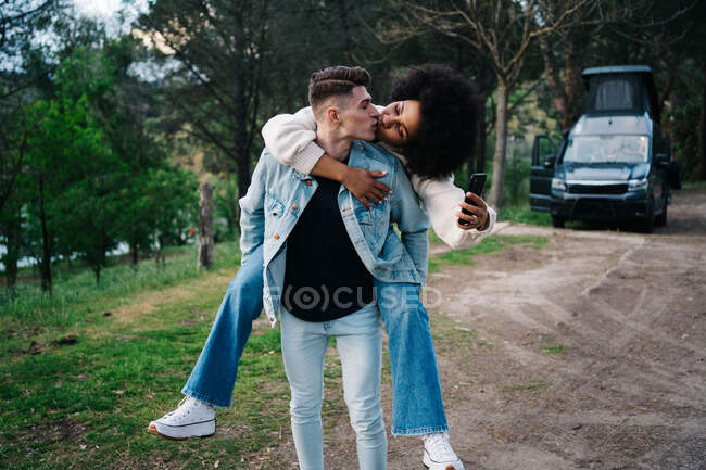Young man giving cheerful ethnic girlfriend piggyback ride against camper and Thai Ridgeback on pathway — Stock Photo