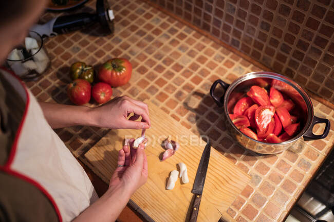 Vista lateral de la hembra étnica en delantal cortando ajo en la tabla de cortar mientras cocina el almuerzo en la cocina en casa - foto de stock