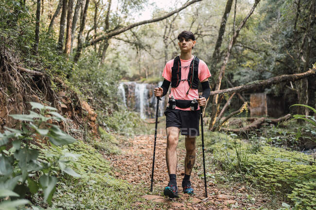 Чоловічий мандрівник з пішохідними полюсами, що йдуть по стежці біля водоспаду в тропічному лісі — стокове фото