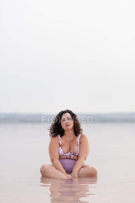 Mujer con curvas graves en bikini sentado en el agua de estanque rosa en verano y mirando a la cámara - foto de stock