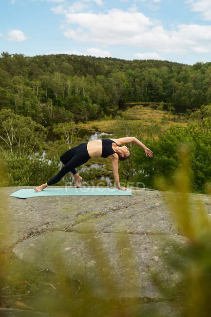Mujer delgada y elegante en leggins negros y sujetador equilibrándose en pose Wild Thing y estirando el cuerpo durante la sesión de yoga contra árboles verdes en el campo - foto de stock