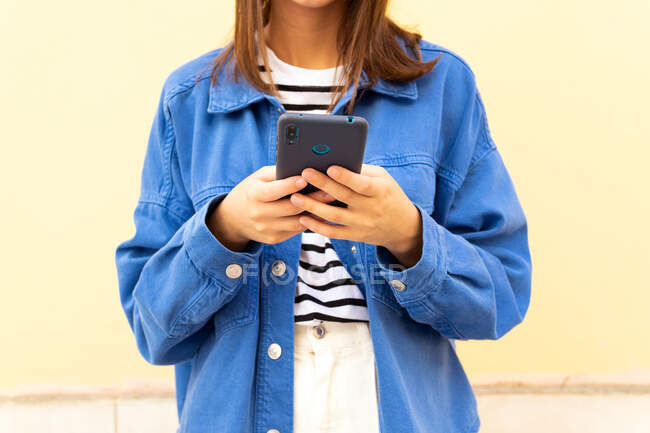 Cortada jovem fêmea irreconhecível na moda mensagens roupa no celular no fundo da parede na rua da cidade e olhando para longe — Fotografia de Stock