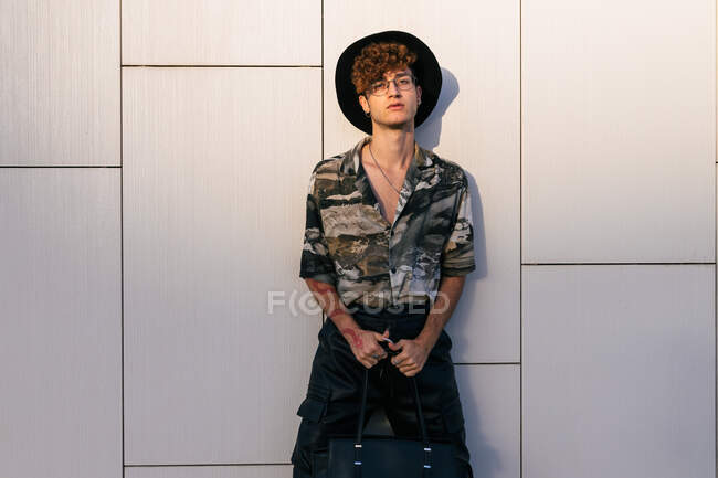 Young vain man in stylish wear with lady's purse standing on tiled wall while looking at camera — Stock Photo