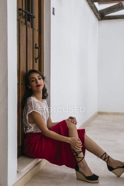 Вид збоку весела етнічна жінка з червоними губами і в модному літньому одязі, що сидить біля дерев'яних дверей у внутрішньому дворику будинку і дивиться на камеру — стокове фото