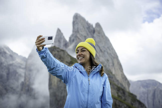 Positiva escursionista femminile in capispalla vibrante scattata su smartphone contro la montagna Seceda durante i viaggi nelle Dolomiti — Foto stock