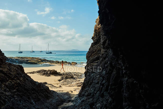Turista femminile in piedi vicino a onde di mare schiumose sulla spiaggia di sabbia bagnata contro scogliera rocciosa e cielo blu nuvoloso durante le vacanze estive a Fuerteventura, Spagna — Foto stock