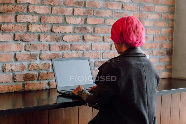Rückseite Ernte Frau mit gefärbten Haaren in stilvollem Mantel sitzt am Tisch in der Nähe Ziegelwand, während mit Laptop — Stockfoto