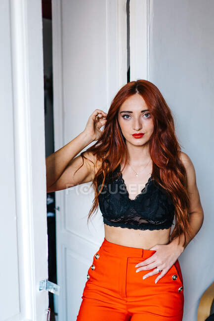 Stylish female with ginger hair and in fashionable outfit leaning on wall near door at home and looking at camera — Stock Photo