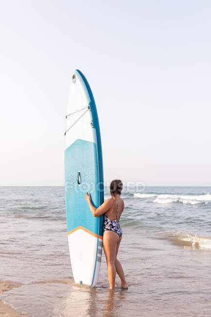 Surfeuse debout avec SUP board bleu sur le bord de mer sablonneux en été et regardant loin — Photo de stock