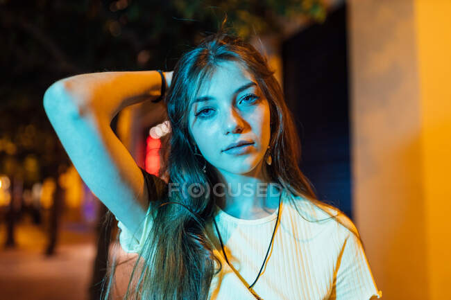 Jeune femme souriante en pendentif aux cheveux longs regardant la caméra sur la route urbaine au crépuscule — Photo de stock
