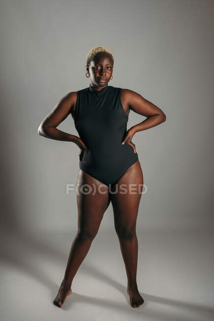 Serious African American curvy female in black bodysuit standing with her hands on her hips on gray background in studio — Stock Photo