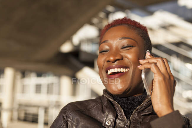 Vista lateral de una hermosa mujer afro negra hablando con su teléfono inteligente mientras sonríe sobre un fondo borroso en un día soleado - foto de stock