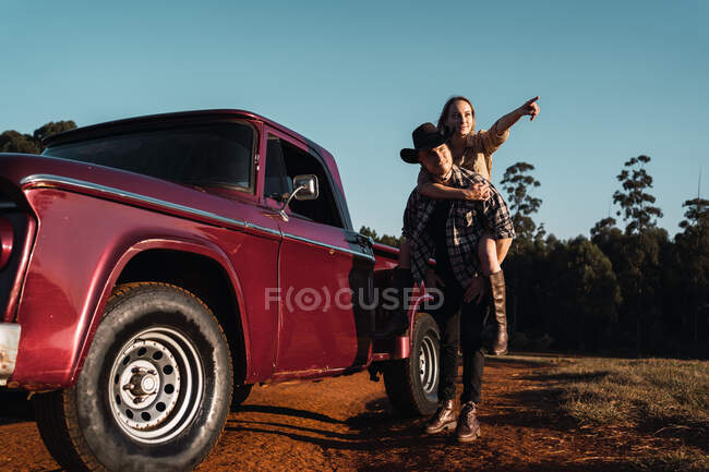 Trendy woman pointing away and piggybacking man standing near vintage pickup car parked on road in nature in evening — Stock Photo