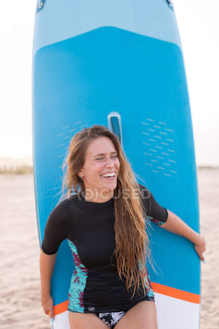 Glückliche Surferin steht im Sommer mit blauem SUP-Board am Sandstrand und schaut weg — Stockfoto