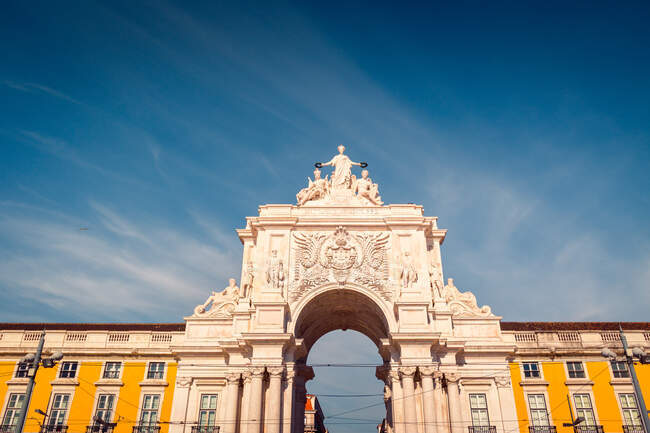 Снизу знаменитая арка Руа Аугуста с классическими орнаментами и скульптурами, расположенная на площади Прака-ду-Комерсио против облачного голубого неба в Лиссабоне, Португалия — стоковое фото
