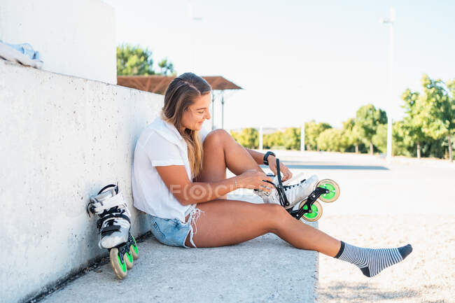 Vista lateral de la alegre patinadora sentada en el suelo en la zona urbana y poniéndose patines mientras disfruta del fin de semana en verano - foto de stock