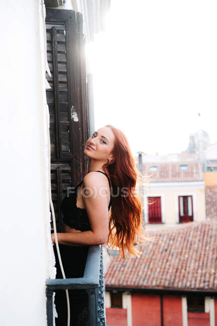 Посміхаючись безтурботна жінка насолоджується сонячним світлом, дивлячись на камеру, стоячи на балконі житлового будинку в місті — стокове фото
