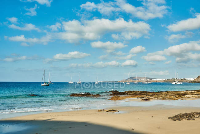 Plage avec voiliers en arrière-plan sur une mer turquoise sous un ciel nuageux par une journée ensoleillée d'été à Fuerteventura, Espagne — Photo de stock