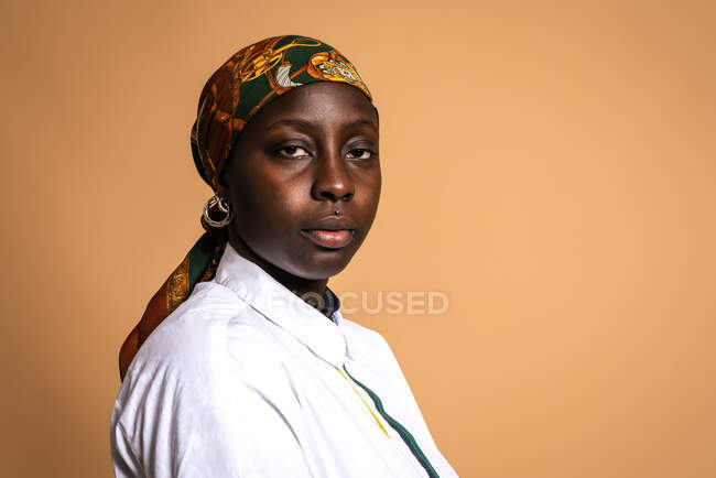 Serious African American female model in trendy headscarf and white shirt looking at camera on beige background in studio — Stock Photo