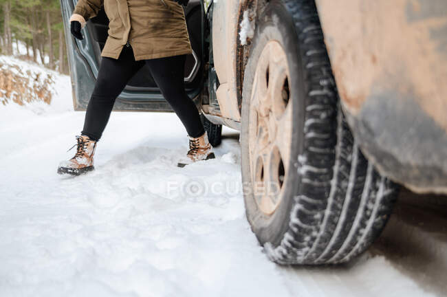 Crop anonymous female in warm clothes getting out of car parked on snowy road in winter woods — Stock Photo