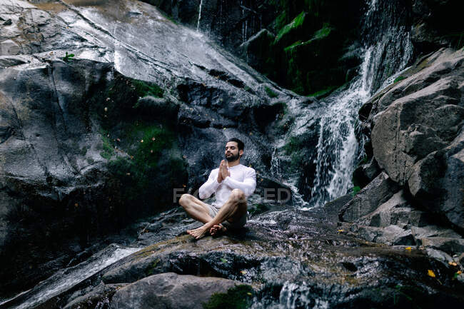 Tranquilo macho sentado em Lótus posar com as mãos de oração na rocha perto de cachoeira e meditar enquanto faz ioga com os olhos fechados — Fotografia de Stock
