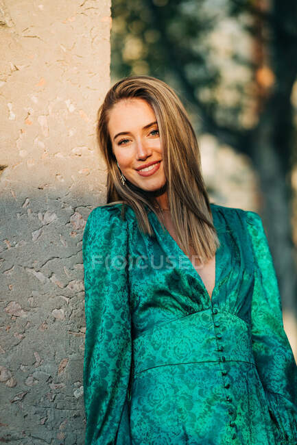 Mulher despreocupada em vestido verde na moda em pé encostado a uma parede na rua e olhando para longe — Fotografia de Stock