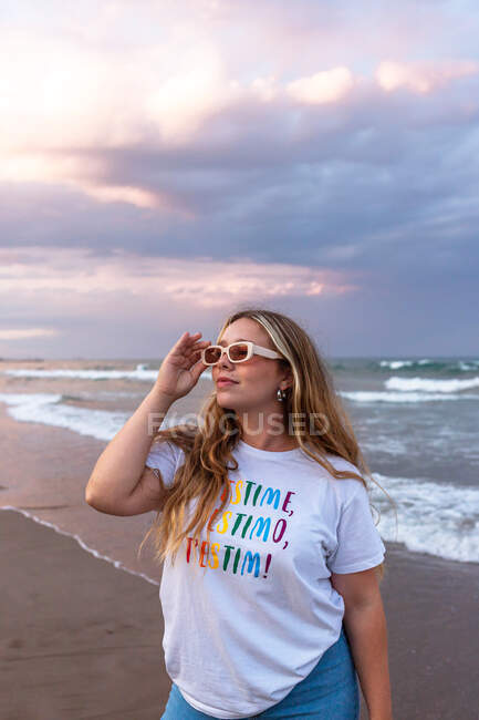 Positive junge Frau mit trendiger Sonnenbrille und stylischem Outfit steht im Sommer abends am Meer — Stockfoto