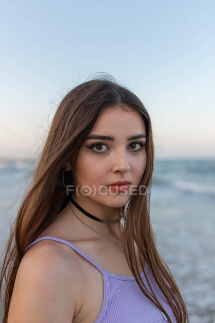 Sonhador jovem fêmea com cabelos longos olhando para a câmera enquanto estava em pé na praia de areia perto do mar ondulando — Fotografia de Stock