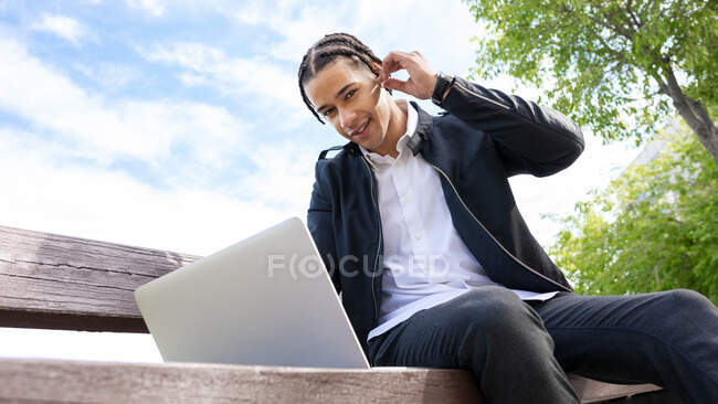 De baixo de freelancer masculino étnico feliz com penteado trançado sentado no banco e digitando no teclado do laptop enquanto trabalhava no projeto remotamente na rua urbana usando fones de ouvido TWS — Fotografia de Stock