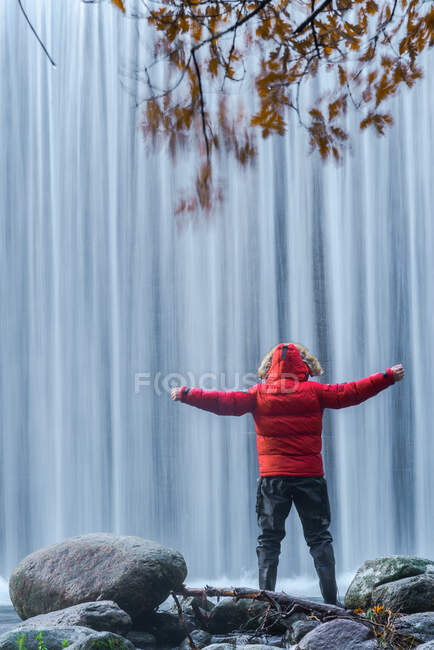 Back view of traveling male in warm clothes standing on boulder with outstretched arms and admiring view of rapid waterfall in long exposure at Lozoya river in Guadarrama National Park — Stock Photo