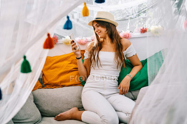 Conteúdo fêmea sentada no sofá e mensagens no celular no dia ensolarado na tenda do quintal — Fotografia de Stock