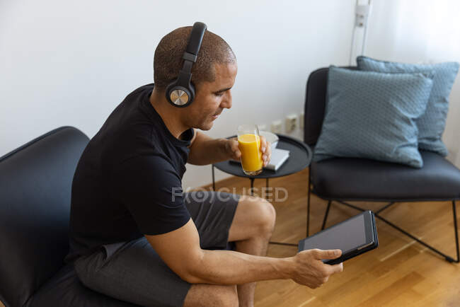 Side view from above of male in headphones watching video on tablet while drinking orange juice and sitting on chair in morning at home — Stock Photo