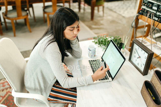 Vista lateral femenina haciendo compra con tarjeta de plástico para la orden durante las compras en línea a través de ordenador portátil - foto de stock