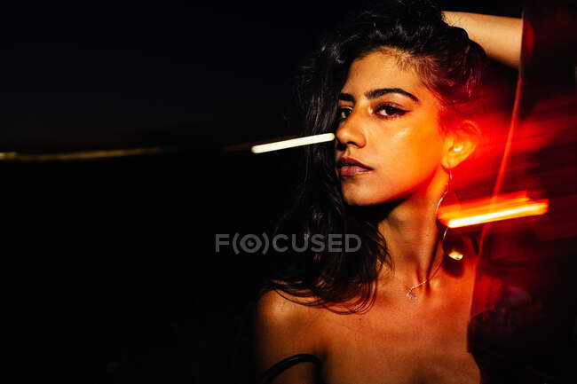 Confident young Hispanic female with long wavy hair and bare shoulders keeping hand on head and looking at camera while standing near glass window with light reflections in darkness at night time — Stock Photo
