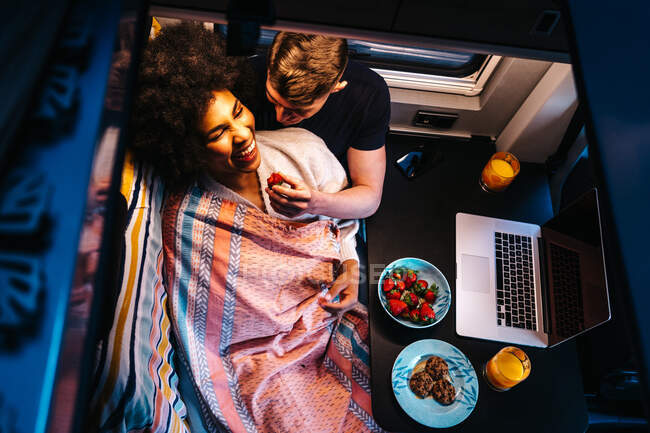 Сверху счастливая многорасовая пара, обедающая рядом с ноутбуком, отдыхающая вместе в кемпере во время отпуска — стоковое фото