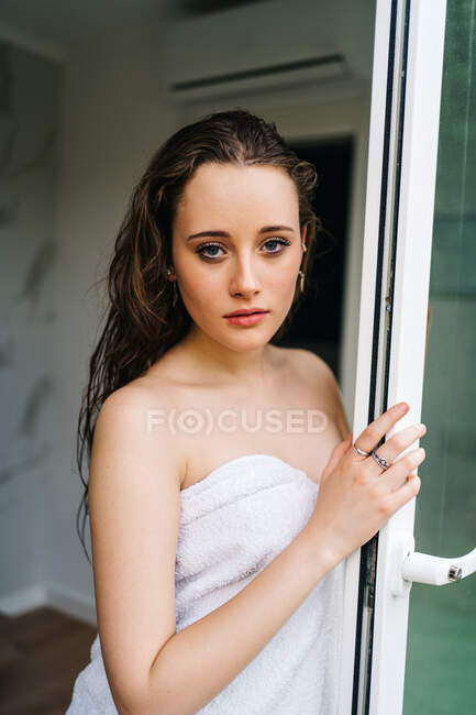 Mujer suave envuelta en toalla blanca de pie con el pelo mojado después de tomar la ducha cerca de la puerta en la terraza y mirando a la cámara - foto de stock