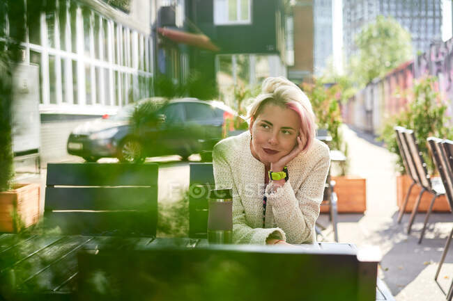 Беззаботная неформальная женщина с окрашенными волосами, сидящая за столом в уличном кафе, опираясь на руку и глядя вдаль во сне — стоковое фото