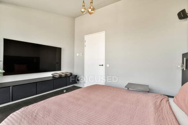 Современный дизайн интерьера светлой спальни с большой кроватью, покрытой розовым одеялом в современном чердачном стиле городской квартиры с большим телевизором — стоковое фото