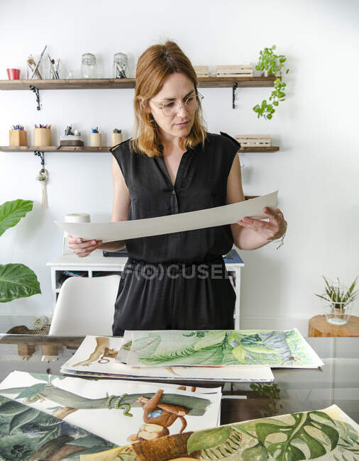 Criativa designer feminina examinando várias pinturas colocadas na mesa e trabalhando no projeto — Fotografia de Stock