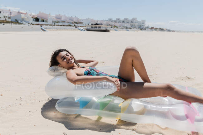 Vista lateral de la mujer feliz en traje de baño acostado en el colchón inflable en la orilla del mar de arena y tomar el sol en el día soleado durante las vacaciones de verano - foto de stock