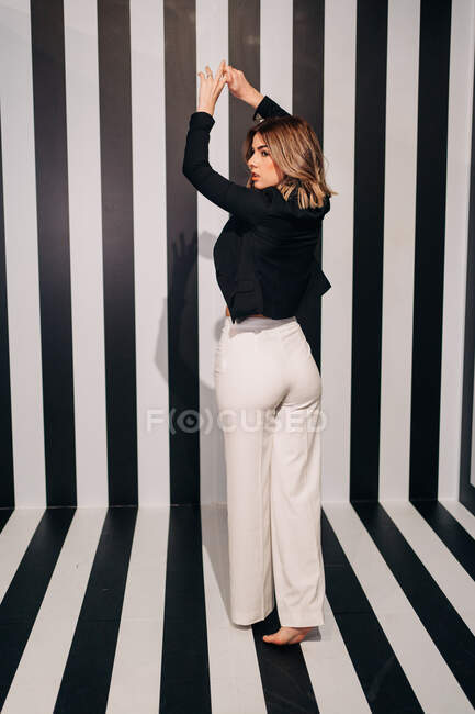 Young seductive woman in white trousers and black jacket looking away on striped floor — Stock Photo