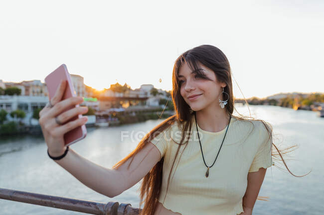 Sincero giovane femmina in ciondolo prendendo autoritratto sul cellulare oltre fiume urbano in serata — Foto stock