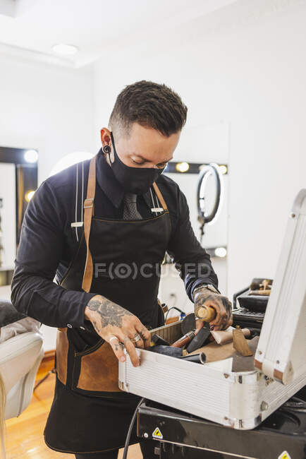 Homem tatuado em máscara pegando vários suprimentos cosméticos da mala enquanto trabalhava no salão de beleza durante a pandemia — Fotografia de Stock