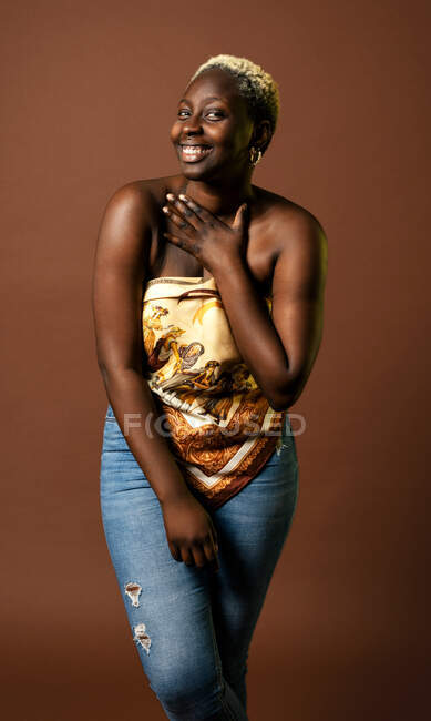 Modelo feminino afro-americano feliz vestindo cachecol de seda top tocando pescoço no fundo marrom em estúdio e olhando para a câmera — Fotografia de Stock