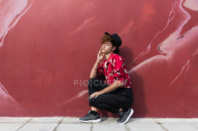 Вид сбоку на модного гомосексуального мужчину с длинными ногтями в декоративной рубашке с закрытыми глазами, приседающего на тротуаре к стене — стоковое фото
