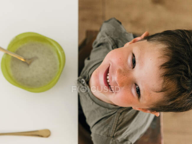 Vista aérea del adorable niño alegre sentado a la mesa a la hora del almuerzo en casa sonriendo a la cámara - foto de stock