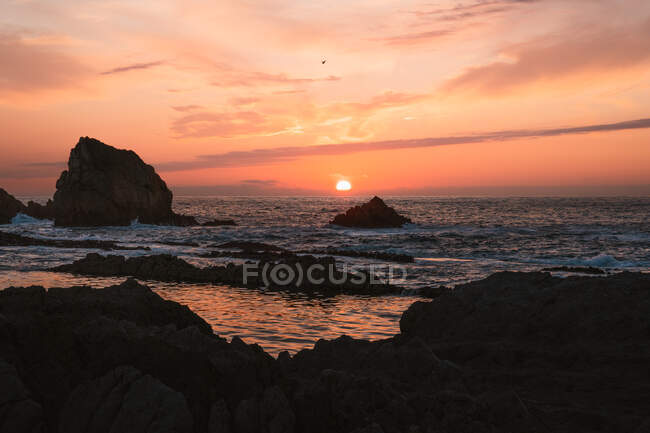 Increíble paisaje tranquilo de puesta de sol sobre el mar ondulado ondulado con rocas bajo el cielo nublado colorido en la noche de verano en Liencres Cantabria España - foto de stock