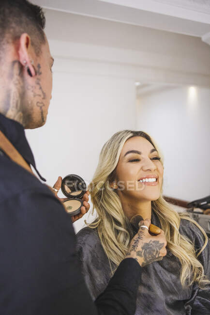 Artista de maquiagem masculina espalhando pó no pescoço de mulher loira otimista durante o trabalho no salão de beleza — Fotografia de Stock